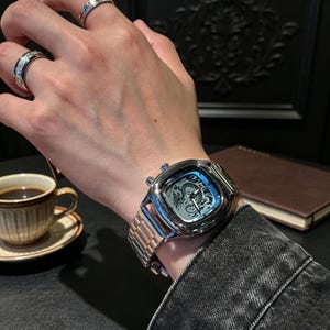 May include: A silver-toned wristwatch with a square case and a metallic link bracelet. The watch face features a detailed black dragon illustration, silver hands, and two side buttons. Two silver rings with embedded stones are worn on the fingers.