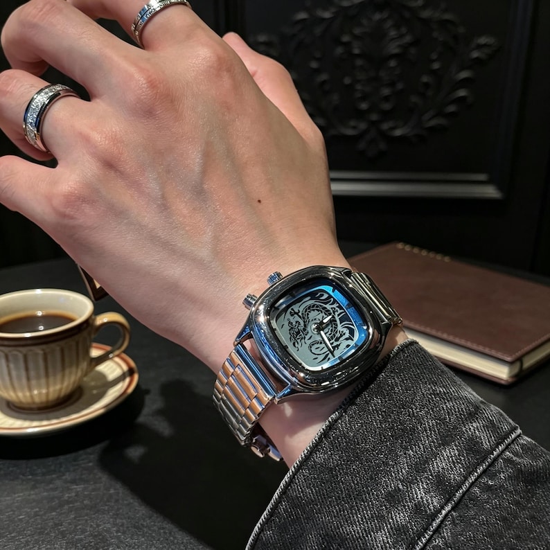 May include: A silver-toned wristwatch with a square case and a metallic link bracelet. The watch face features a detailed black dragon illustration, silver hands, and two side buttons. Two silver rings with embedded stones are worn on the fingers.