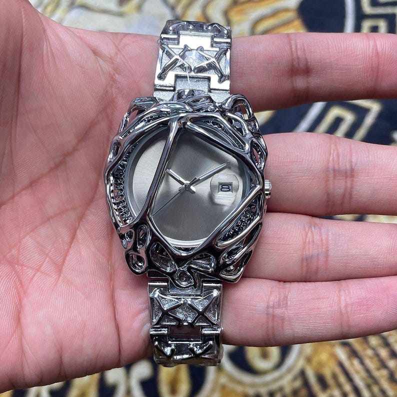 May include: A silver-toned wristwatch with an intricate, openwork design around the face and band. The watch face is a light gray with silver hands and a date display. The band features geometric patterns.