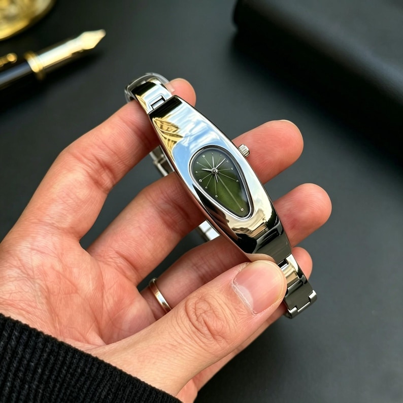 以下が含まれることがあります： 楕円形の緑色の文字盤とシルバーのバンドが付いた、シルバーカラーの腕時計。この時計は、ユニークな細長いデザインと洗練された仕上げが特徴です。手で持たれています。