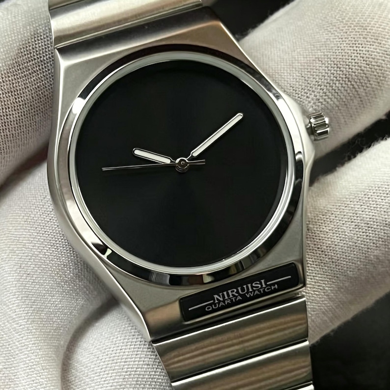 May include: A stainless steel wristwatch with a black face and silver hands. The watch has a minimalist design with the text "NIRUISI QUARTA WATCH" on the band. The watch is on a silver metal band.