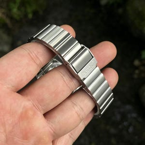 May include: A silver-colored watch band with a folding clasp. The band is made of multiple rectangular links. The clasp is engraved with the word "ZENGGE". The band is held in a hand.