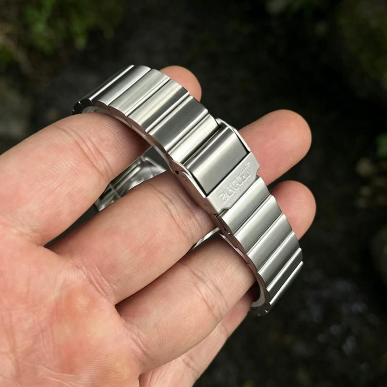 May include: A silver-colored watch band with a folding clasp. The band is made of multiple rectangular links. The clasp is engraved with the word "ZENGGE". The band is held in a hand.