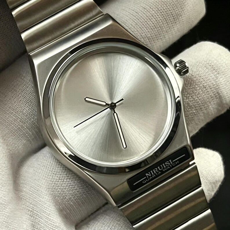 May include: A silver-toned wristwatch with a minimalist design. The watch features a circular face with a brushed metal finish, black hands, and a rectangular case. The watch band is made of matching silver metal links. The text "NIRUISI QUARTA WATCH" is printed on the watch.