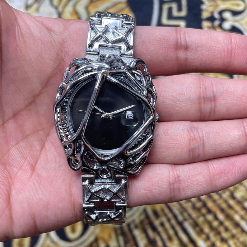 May include: Silver-toned wristwatch with a black watch face and a unique, cage-like design around the dial. The watch band features geometric patterns. The watch displays the date. A stylish accessory.