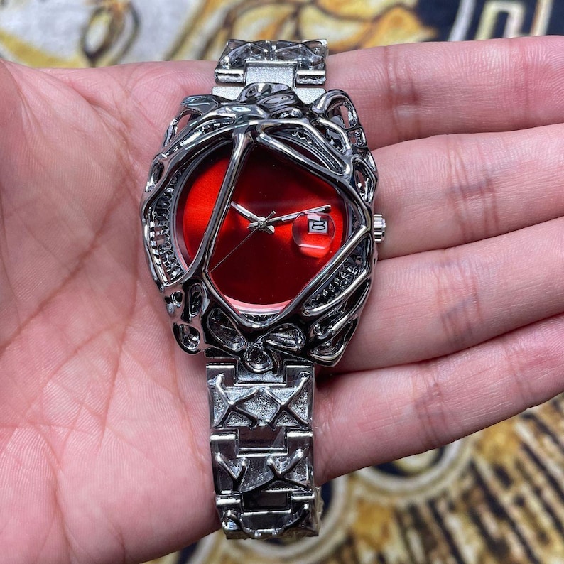 May include: A silver-toned wristwatch with a red face and a date display. The watch features an intricate, openwork design around the face and on the band. The watch band is made of interlocking geometric shapes.