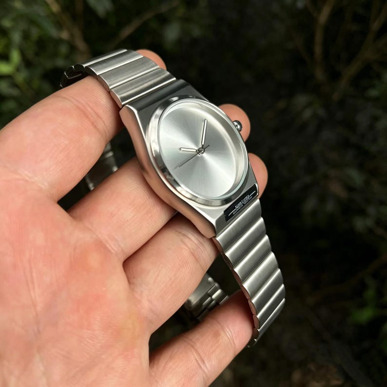May include: A silver-toned wristwatch with a minimalist design. The watch features a round face with a brushed metal finish and a matching metal band. The watch is held in a hand against a blurred green background.
