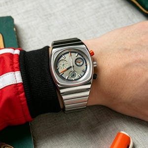 May include: A silver-toned wristwatch with a square face and a matching metal band. The watch face is gray with blue and orange accents, and two smaller dials. The watch is on a wrist with a red and black sleeve.