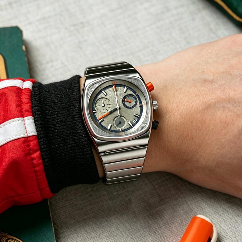 May include: A silver-toned wristwatch with a square face and a matching metal band. The watch face is gray with blue and orange accents, and two smaller dials. The watch is on a wrist with a red and black sleeve.