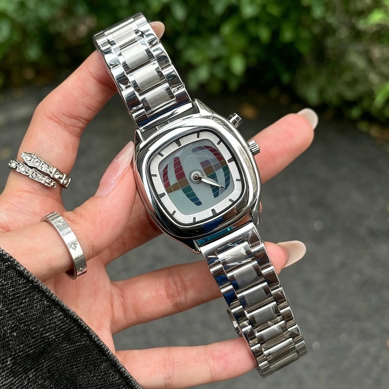 Puede incluir: Un reloj de pulsera plateado con una esfera cuadrada y una correa de metal a juego. La esfera del reloj presenta un dise&ntilde;o abstracto y colorido. El reloj se muestra en una mu&ntilde;eca, mostrando su acabado pulido y dise&ntilde;o moderno.