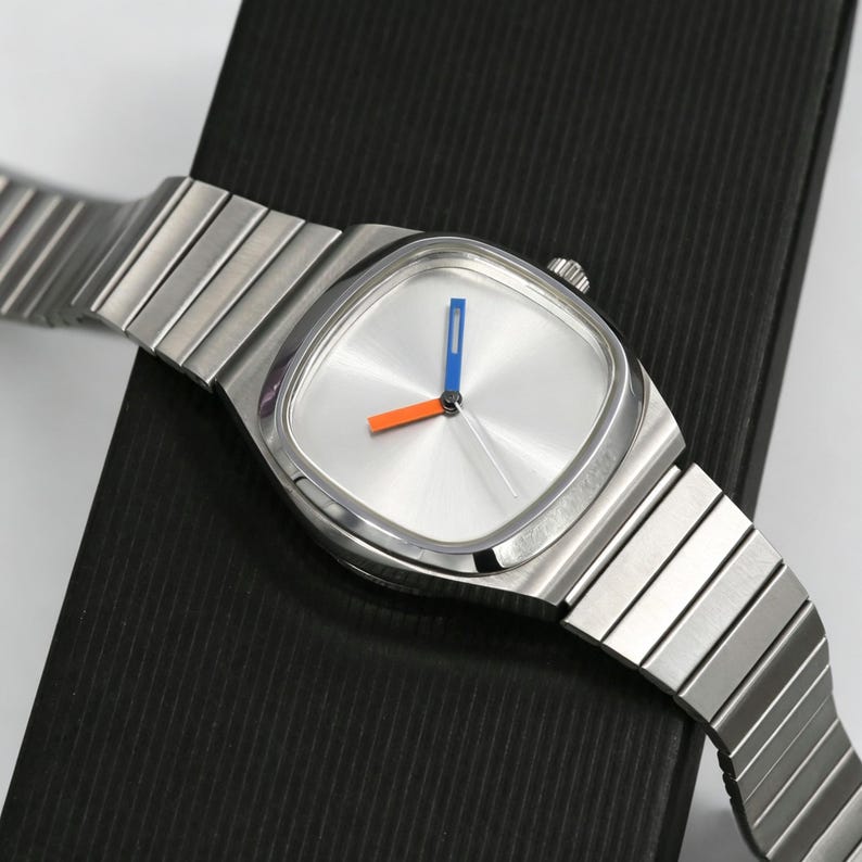 May include: A silver-toned wristwatch with a square face and a matching metal band. The watch face is silver with blue and orange hands. The watch is resting on a black surface.