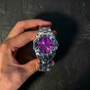 May include: A silver-toned wristwatch with a unique, textured design. The watch face is a vibrant purple, and the band is made of linked metal segments. The watch is held in a hand against a dark background.