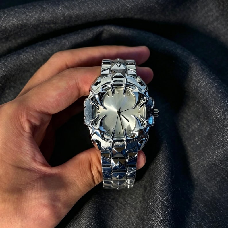 May include: A silver-toned wristwatch with a unique, ornate design. The watch features a textured, geometric band and a face with a stylized, floral-like pattern. The watch is held in a hand against a dark background.