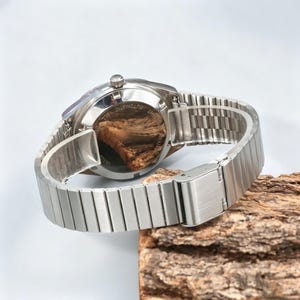May include: A silver-toned wristwatch with a metal band. The watch is displayed on a piece of wood. The watch face is visible, and the back of the watch is also visible, showing the inner workings.