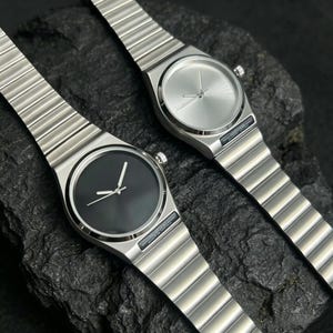May include: Two minimalist stainless steel watches with matching metal bands. One watch has a black face, the other a silver face. Both have silver hands and a small crown. The watches are displayed on a dark, textured surface.