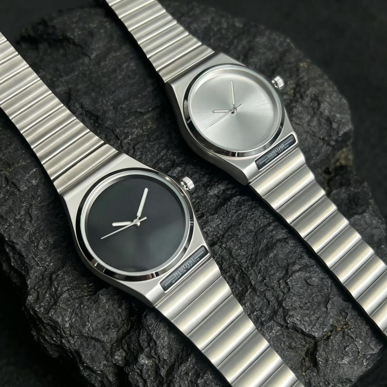 May include: Two minimalist stainless steel watches with matching metal bands. One watch has a black face, the other a silver face. Both have silver hands and a small crown. The watches are displayed on a dark, textured surface.