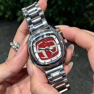 Puede incluir: Un reloj de pulsera de acero inoxidable con una esfera roja cuadrada con un dise&ntilde;o gr&aacute;fico blanco. El reloj tiene una correa plateada y una caja plateada. El reloj est&aacute; siendo sostenido por una persona.
