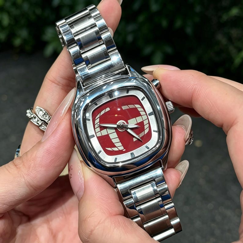 Puede incluir: Un reloj de pulsera de acero inoxidable con una esfera roja cuadrada con un dise&ntilde;o gr&aacute;fico blanco. El reloj tiene una correa plateada y una caja plateada. El reloj est&aacute; siendo sostenido por una persona.