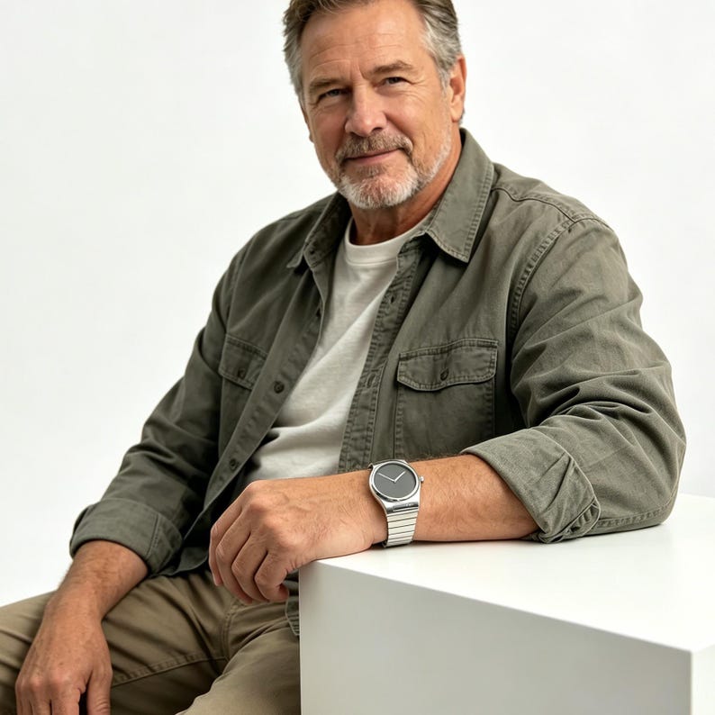 May include: A silver wristwatch with a black face is worn on a wrist. The watch has a metallic band. The person is wearing a green shirt and khaki pants. The watch is a stylish accessory.