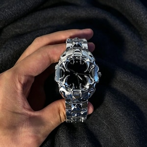 May include: A silver-toned wristwatch with a black watch face. The watch has a unique, ornate design with a geometric pattern on the band. The watch is held against a dark gray background.