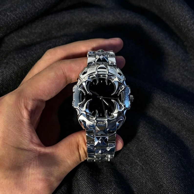May include: A silver-toned wristwatch with a black watch face. The watch has a unique, ornate design with a geometric pattern on the band. The watch is held against a dark gray background.