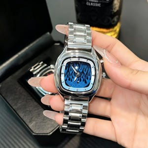 May include: A silver-toned wristwatch featuring a square-shaped case with rounded corners and a metallic link bracelet. The watch face displays a vibrant blue flame pattern with silver hands and hour markers, housed in a polished metal frame.