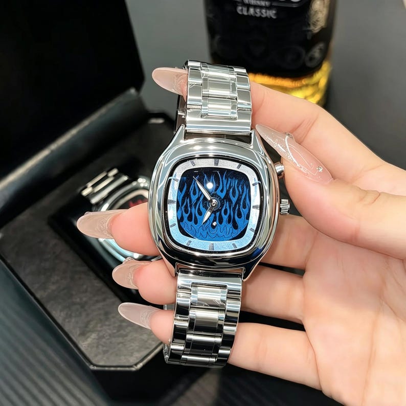 May include: A silver-toned wristwatch featuring a square-shaped case with rounded corners and a metallic link bracelet. The watch face displays a vibrant blue flame pattern with silver hands and hour markers, housed in a polished metal frame.