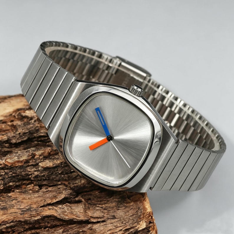 May include: A silver-toned wristwatch with a square face and a silver band. The watch face has a radial brushed pattern with blue and orange hands. The watch is displayed on a piece of wood.