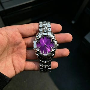 May include: A silver-toned wristwatch with a unique design. The watch features a vibrant purple face and a detailed, textured band. The watch's face has a distinctive shape, and the band is made of interconnected links.