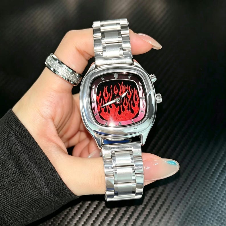 May include: A silver-tone analog wristwatch featuring a square-shaped case with rounded corners and a vibrant red flame graphic on the dial. The watch includes a polished metal link bracelet, silver hands, and two side buttons on the right side.