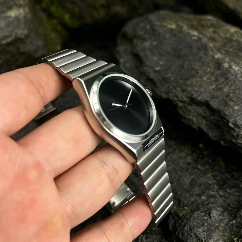 May include: A silver wristwatch with a black watch face. The watch has a sleek, minimalist design with a metal band. The hands are white, and the watch is held in a person's hand.