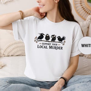 May include: A white short-sleeved t-shirt featuring a graphic of four black crows perched on a branch with the text "SUPPORT YOUR LOCAL MURDER" printed in black lettering below the birds. A circular white label on the right reads "WHITE".