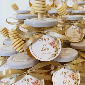 Peut inclure: Petits pots de miel en verre, chacun surmonté d'un bâtonnet à miel en bois et d'un couvercle blanc. Les pots sont ornés de rubans dorés et d'étiquettes indiquant "Aya & Léo 17 Janvier 2026".