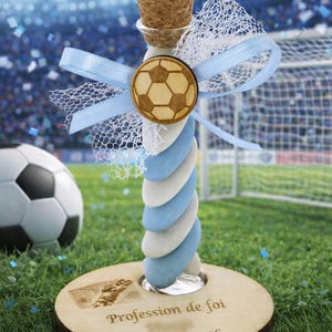 Peut inclure: Une bouteille décorative en verre avec un bouchon en liège, ornée d'un ballon de football en bois et d'un ruban bleu clair. La bouteille repose sur une base en bois avec le texte "Profession de foi" et la date "20 avril 2026".
