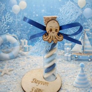 Peut inclure: Un tube en verre rempli de bonbons bleus et blancs, surmonté d'un charm en bois représentant une pieuvre et d'un ruban bleu. Le tube repose sur une base en bois avec le texte "Dayvon 5 ans". Thème anniversaire.