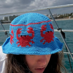 May include: A bright blue crochet bucket hat featuring a repeating pattern of red crabs around the crown, separated by a thin red horizontal line. The hat has a flexible, downward-sloping brim and a textured, hand-stitched appearance.