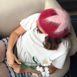 May include: A person wearing a two-tone pink and red fuzzy hat and a white t-shirt. The person is holding a handmade crochet daisy flower with white petals and a yellow center. The person is wearing blue and white patterned shorts.