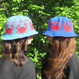 May include: Two blue crochet bucket hats with red crab designs. The hats have a wide brim and are worn by two people. The background is a lush green foliage.