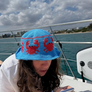 Puede incluir: Un sombrero de pescador azul tejido a ganchillo con diseños de cangrejos rojos. El sombrero tiene un ala ancha. El fondo muestra el mar y un cielo nublado. Un accesorio de verano.