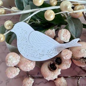 以下が含まれることがあります： 白い紙の鳥の切り抜きで、花のディテールがあり、ピンクのキラキラしたベリーとクリーム色の真珠のような装飾品の中にあります。緑の葉と枝が含まれており、繊細で装飾的なシーンを作り出しています。
