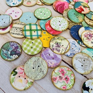 May include: A collection of decorative, round buttons with various floral, patterned, and vintage designs. The buttons feature a variety of colors, including pink, green, blue, and yellow. Each button has two holes for sewing and a distressed, aged appearance.