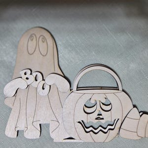 May include: Wooden Halloween decoration featuring a ghost with the word "Boo" and a jack-o'-lantern with a handle and candy corn. The ghost has large eyes and a draped sheet. The jack-o'-lantern has a carved face. The decoration is made of light-colored wood.