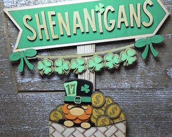 May include: Wooden St. Patrick's Day decoration featuring a green arrow sign with the word "SHENANIGANS" and a banner of shamrocks. A leprechaun figure sits in a basket filled with gold coins. The number "17" is on the leprechaun's hat.