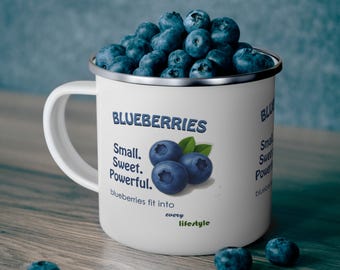 Może przedstawiać: Biały emaliowany kubek wypełniony świeżymi jagodami. Kubek ma srebrną obwódkę i biały uchwyt. Tekst na kubku brzmi "BLUEBERRIES" i "Small. Sweet. Powerful." z ilustracją jagody. Jagody są rozrzucone na drewnianej powierzchni.