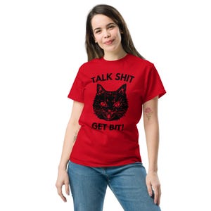 May include: A red t-shirt featuring the text "TALK SHIT GET BIT!" above a black cat graphic. The cat has an angry expression. The shirt is made of a soft material and is being worn.