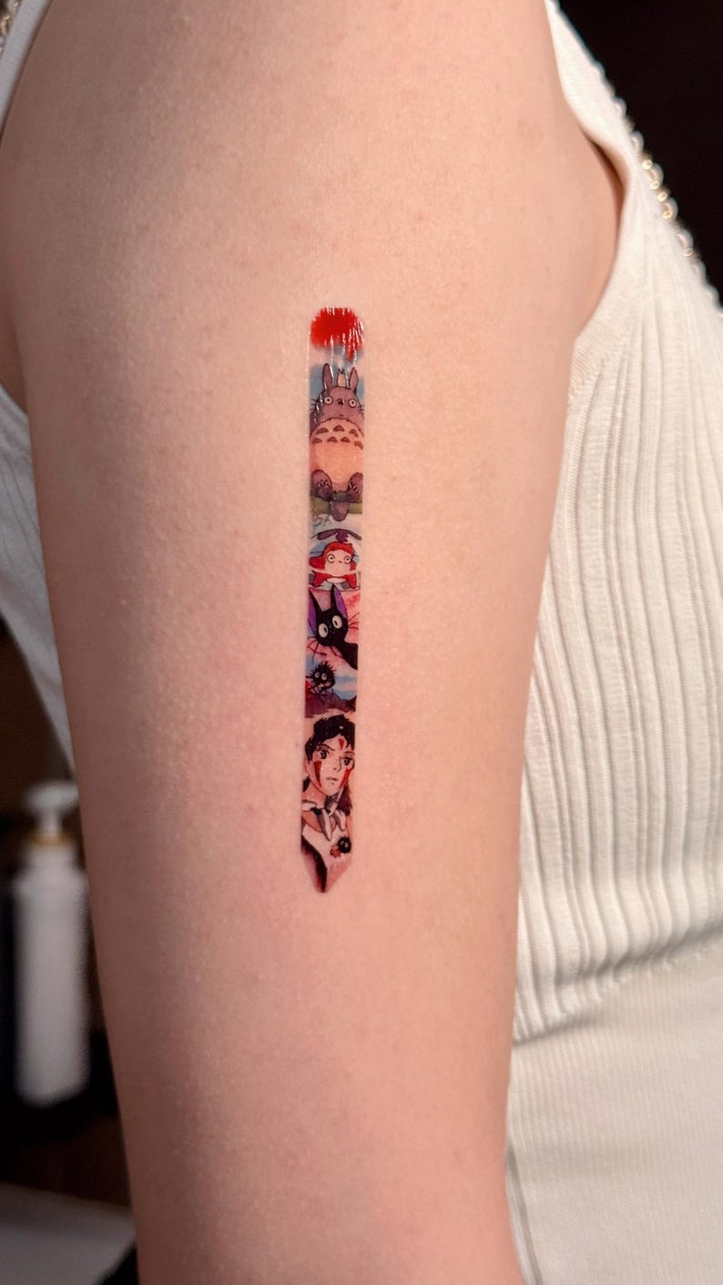 K&ouml;nnte beinhalten: Ein vertikales Tattoo auf dem Arm einer Person zeigt eine Reihe von Figuren aus dem Animationsfilm 'Mein Nachbar Totoro'. Zu den Figuren geh&ouml;ren Totoro, der Katzenbus und ein junges M&auml;dchen.