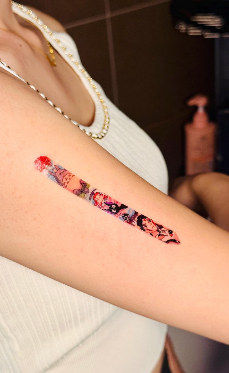 K&ouml;nnte beinhalten: Ein farbenfrohes tempor&auml;res Tattoo auf dem Arm einer Person. Das Tattoo zeigt verschiedene Figuren aus beliebten Anime- und Manga-Serien, darunter Totoro, Kiki liefert aus und Studio Ghibli.