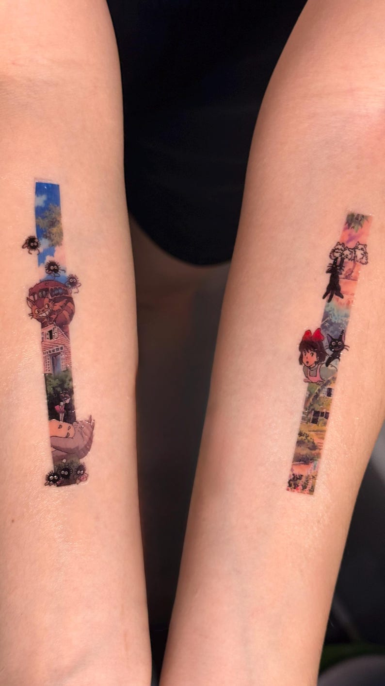 K&ouml;nnte beinhalten: Zwei Tattoos auf den Armen einer Person. Der linke Arm hat ein Tattoo einer Szene aus dem Film "Howls bewegliches Schloss" mit dem Schloss, einer schwarzen Katze und einem M&auml;dchen. Der rechte Arm hat ein Tattoo einer Szene aus dem Film "Kikis kleiner Lieferservice" mit Kiki und ihrer schwarzen Katze Jiji.