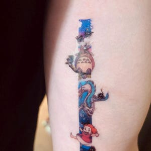 K&ouml;nnte beinhalten: Ein farbenfrohes Tattoo auf dem Arm einer Person mit Figuren aus dem Animationsfilm 'Mein Nachbar Totoro'. Das Tattoo zeigt Totoro, den Katzenbus und ein junges M&auml;dchen.