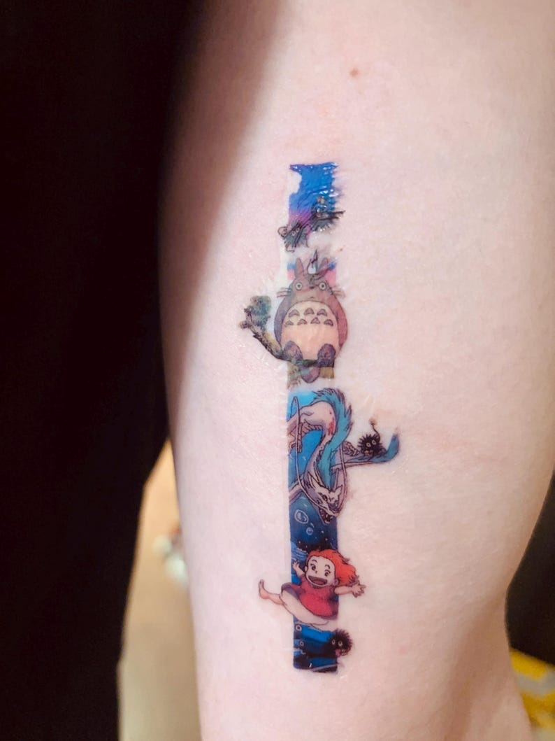 K&ouml;nnte beinhalten: Ein farbenfrohes Tattoo auf dem Arm einer Person mit Figuren aus dem Animationsfilm 'Mein Nachbar Totoro'. Das Tattoo zeigt Totoro, den Katzenbus und ein junges M&auml;dchen.